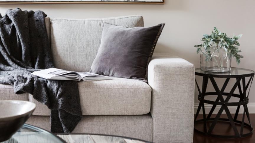 A light grey sofa in a neutrally decorated room with a dark grey blanket draped over and a book open on the sofa