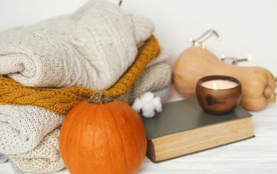 A collection of knitted jumpers stacked next to a pumpkin, a butternut squash, a book and a lit candle