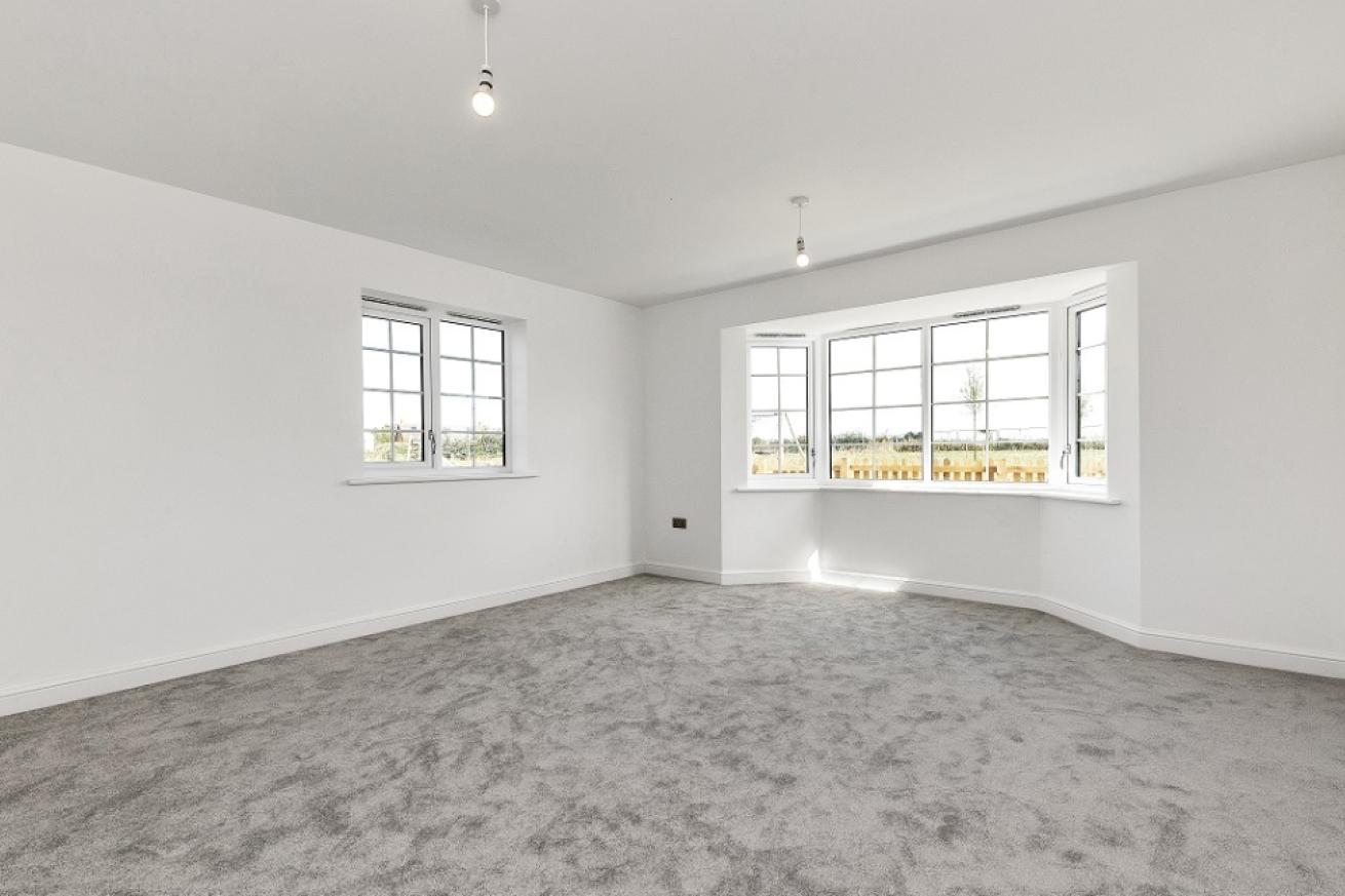 A spacious empty bedroom which is decorated all white with a grey carpet with a bay window looking into the garden