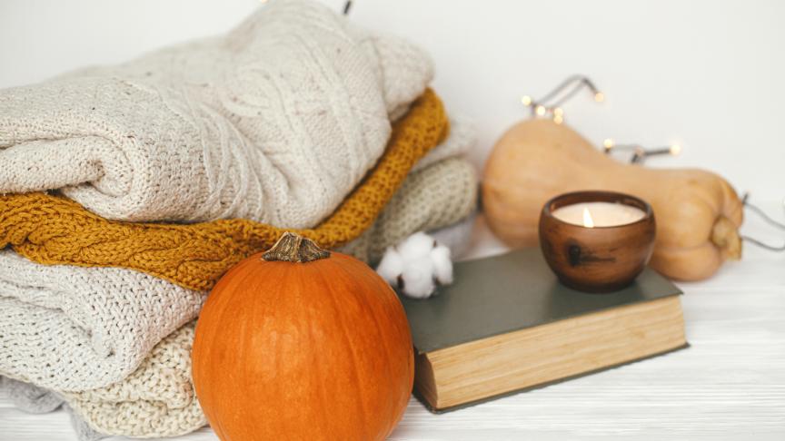 A collection of knitted jumpers stacked next to a pumpkin, a butternut squash, a book and a lit candle