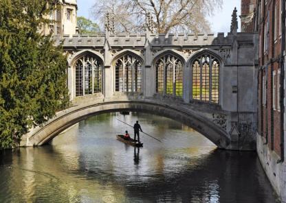 A person standing on a boat punting down the river in Cambridge about to go under a beautiful historic bridge