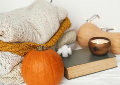 A collection of knitted jumpers stacked next to a pumpkin, a butternut squash, a book and a lit candle